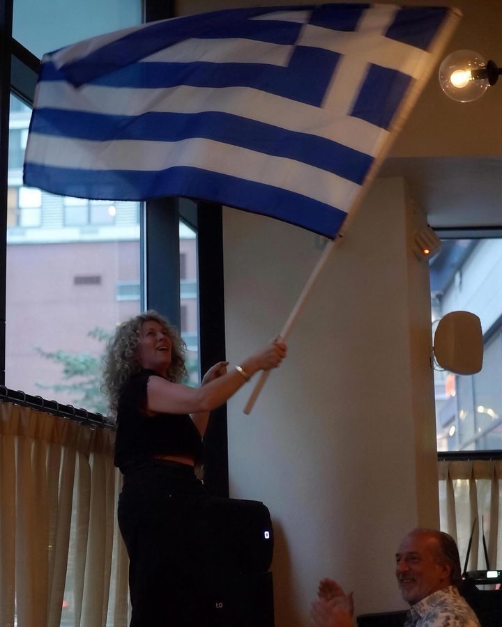 A guest waving the Greek flag as the room cheers during Greek Night at IKOS
